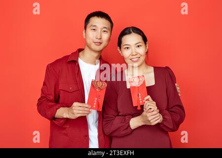 Waist up portrait of young Chinese couple holding red hongbao envelopes with good luck inscription and smiling at camera, copy space Have overflowing abundance every year Stock Photo
