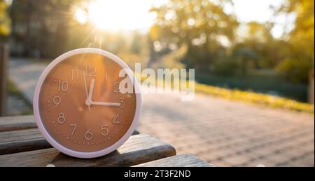 The clock stands on a bench in the park on a sunny autumn day. Change time concept. Planning your day and managing time. Autumn fall season. DST. Sele Stock Photo