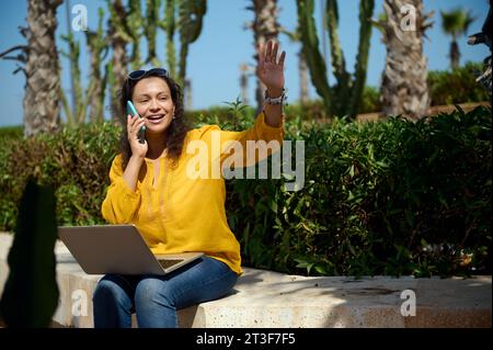 Smiling multitasking busy happy young adult business woman, in casual wear, waving hand greeting someone, talking on smart mobile phone, online workin Stock Photo