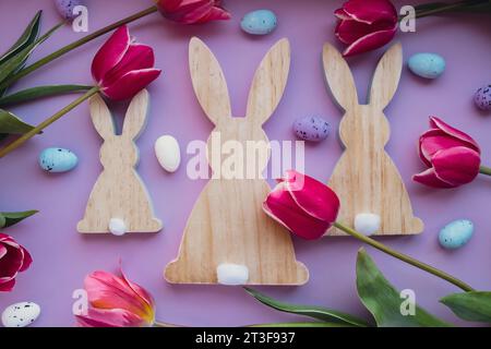 Easter eggs and wooden bunnies with pink tulips on violet pastel background. Flat lay composition, top view. Decoration minimalist modern design template. Floral layout Concept of creative spring holiday mock up.  Stock Photo