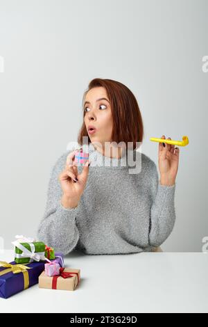 shocked woman in sweater holding tiny Christmas gift and yellow party horn on grey backdrop Stock Photo