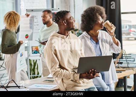 joyous team members with coffee and laptop in hands with their friends on backdrop, working process Stock Photo