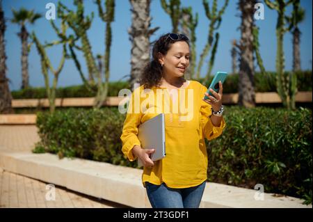 Middle Eastern confident beautiful female entrepreneur, business woman holding a laptop, checking mobile application on her modern smartphone, strolli Stock Photo