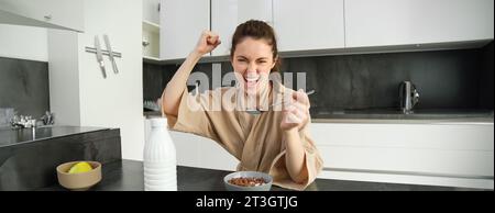 Portrait of enthusiastic young woman eating cereals with milk, looking excited and happy, sitting near kitchen worktop and having breakfast, raising Stock Photo