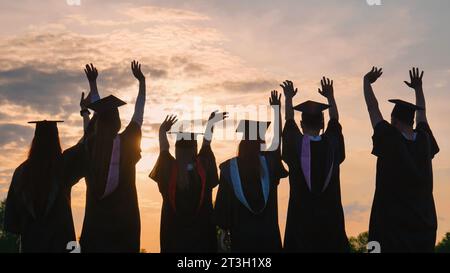 Silhouettes of graduates in black robes wave their arms against the evening sunset and toss their caps. Stock Photo