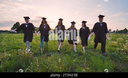 Six graduates in robes walking through a meadow in the evening. Stock Photo