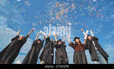 Happy graduates throwing up colorful confetti against the blue summer sky. Stock Photo