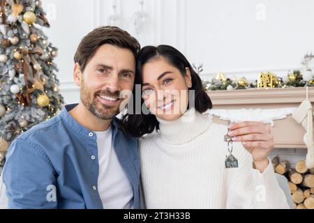 Close-up portrait of young man and woman, happy family and loving couple, sitting hugging at home on sofa near Christmas tree and smiling at camera. Stock Photo