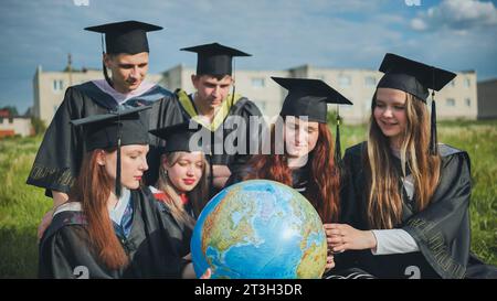 Graduates in black robes examine a geographical globe sitting on the grass. Stock Photo