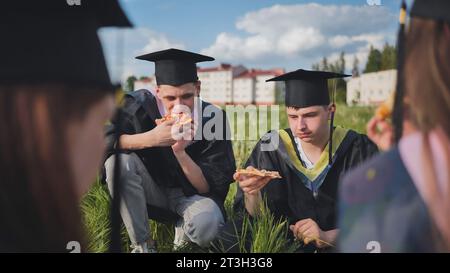 Graduates in black suits eating pizza in a city meadow. Stock Photo