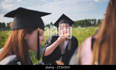 Graduates in black suits eating pizza in a city meadow. Stock Photo