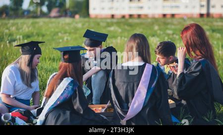 Graduates in black suits eating pizza in a city meadow. Stock Photo
