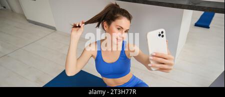 Portrait of beautiful female athlete, woman doing workout at home on rubber yoga mat, takes selfie on smartphone, makes photos for social media of Stock Photo