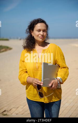 Confident portrait of a beautiful multi-ethnic woman with laptop, looking at camera, standing in the square, dressed in casual yellow shirt and blue d Stock Photo