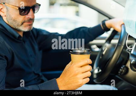 man drinking takeaway coffee inside the car with a protective mask hanging from the rearview mirror of the car. Stock Photo