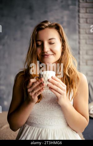With a delightful smile, the young lady enjoys her coffee, appreciating the morning bliss. This image encapsulates the tranquil ambiance of morning co Stock Photo