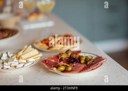 Two glasses of cool champagne and plates with various snacks on a table. White plates with slices of ham, salami, capers, olives and cheese. Cozy inte Stock Photo