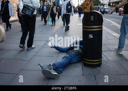 Homeless man sprawled across the pavement along Oxford Street holding a plastic cup in hope to be given change by pedestrians passing by, London, UK Stock Photo