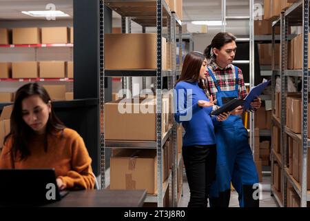 Storehouse managers comparing orders checklist on clipboard and digital tablet. Warehouse asian employees standing near shelf full of cardboard boxes and discussing inventory report Stock Photo