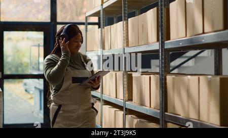 Joyful warehouse chief supply chain officer listening music while inspecting merchandise. Cheerful african american logistics coordinator wearing heaphones while walking through repository rows Stock Photo