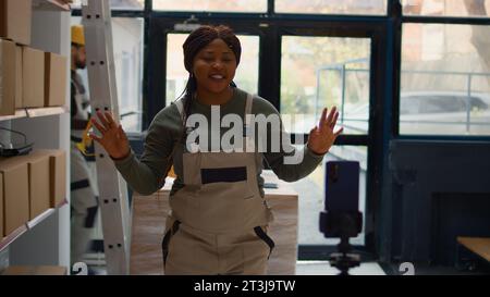 Warehouse chief supply chain officer using smartphone to make training video for interns. African american supervisor films herself in depository showing trainees how to seal cardboard boxes Stock Photo