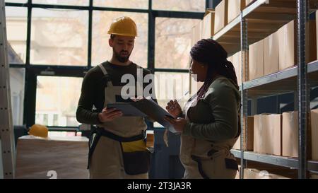 Logistics coordinators in warehouse inputing updated products info on online shopping website, walking through retail workspace aisles analyzing labels on wares, crosschecking with inventory list Stock Photo
