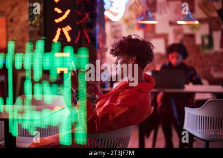 Side view of young man in bomber jacket scrolling on tablet screen while sitting in cyberpunk bar or cafe with neon lights and vibrant colors Stock Photo