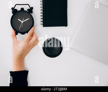 Close up person s hand holding black alarm clock spiral notepad laptop takeaway coffee cup white desk Stock Photo