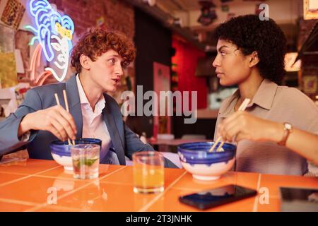 Two young intercultural colleagues or dates having chat at lunch break in Japanese cafe or restaurant while eating ramen and looking at one another Stock Photo