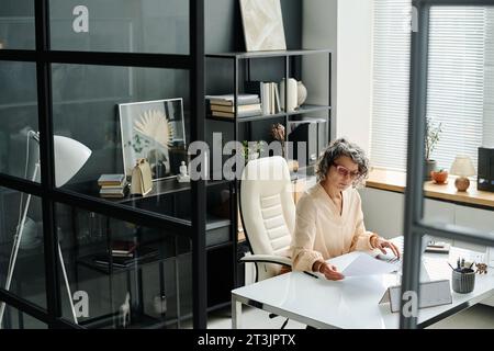 Mature female boss with financial paper sitting by workplace in office and looking through points of contract while checking data before entering Stock Photo