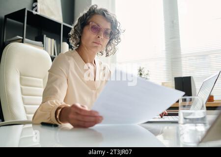 Focus on mature female chief executive officer reading financial paper while sitting by workplace in front of camera and checking data Stock Photo