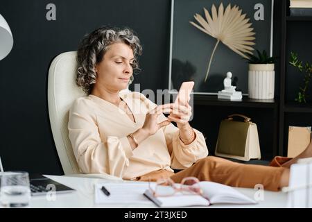 Mature relaxed female ceo in quiet luxury attire using mobile phone while sitting by her workplace in office and keeping legs on desk Stock Photo