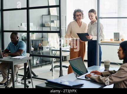 Experienced female boss and her young subordinate discussing document while standing in front of two intercultural employees with laptops Stock Photo