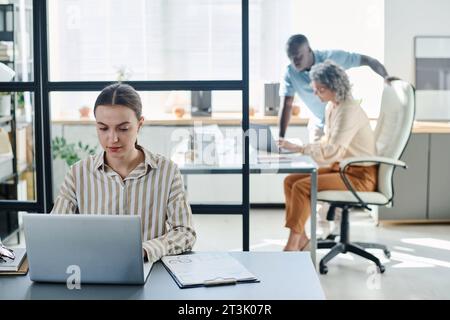 Young secretary or analyst using laptop while sitting by desk with financial document with graphic data and looking through information on screen Stock Photo