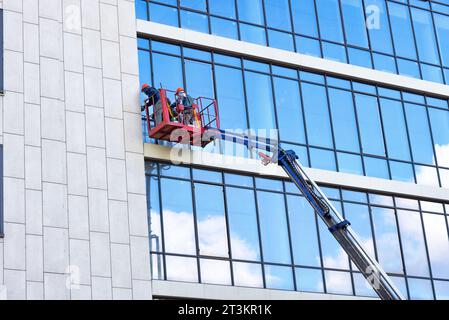 External glass facade window washers, while in a construction lifting cradle, clean the glass facade of a building on a clear day. Stock Photo