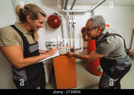 Team of heating engineers discussing and checking a old gas heating system, holding a Clipboard with checklist at a boiler room in a house. Gas heater Stock Photo