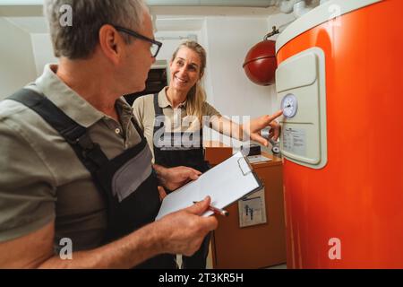 Team of heating engineers checks a old gas heating system thermostat, holding a Clipboard with checklist at a boiler room in a house. Gas heater repla Stock Photo