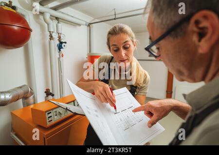 Team of heating engineers checks a old gas heating system with a paper instruction at a boiler room in a house. Gas heater replacement obligation conc Stock Photo