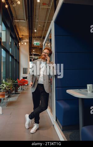 Smiling entrepreneur with laptop is talking phone standing in modern office near window Stock Photo