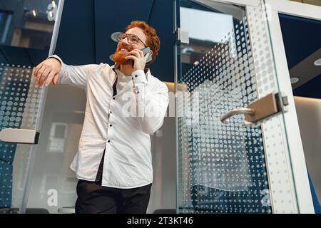 Bearded businessman in eyeglasses is talking phone standing in modern coworking space Stock Photo