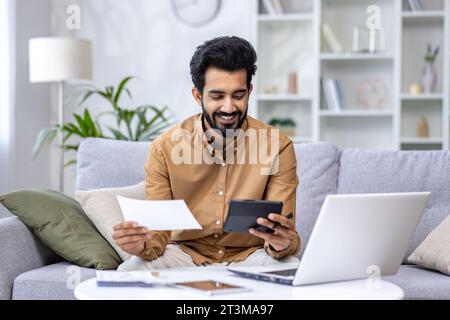 Young man working at home with documents and papers, homework, paying bills and loans, hispanic sitting on sofa in living room, using laptop and calculator. Stock Photo