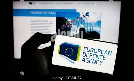 Person holding cellphone with logo of EU institution European Defence Agency (EDA) in front of webpage. Focus on phone display. Stock Photo
