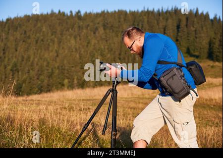 Professional photographer taking pictures in mountains. Man focusing his camera to take nice photo. Side view of young male adjusting camera on background of hills. Stock Photo