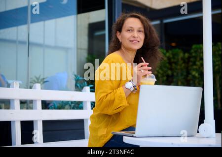 Happy smiling successful middle aged African American woman, business lady, freelance entrepreneur drinking freshly squeezed juice and working online Stock Photo