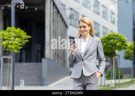 Successful woman boss walks outside office building outdoors, in business suit, businesswoman uses app on phone, smiles contentedly reads internet pages, browses news, writes text message, call. Stock Photo