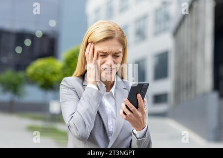 Close-up of upset senior business woman holding phone, female worker in business suit receiving bad accusations online, boss using app on smartphone, walking outside office building in city. Stock Photo