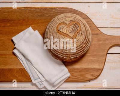 Flat lay of a gluten-free sourdough boule scored with a heart shape, placed on a wooden breadboard. Stock Photo
