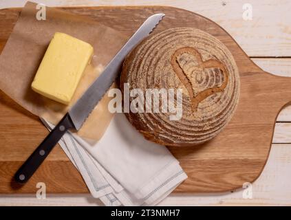 Flat lay of a gluten-free sourdough boule scored with a heart shape, placed on a wooden breadboard with a block of butter. Stock Photo