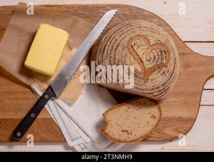 Flat lay of a gluten-free sourdough boule scored with a heart shape, placed on a wooden chopping board with a block of butter and a bread knife. Stock Photo