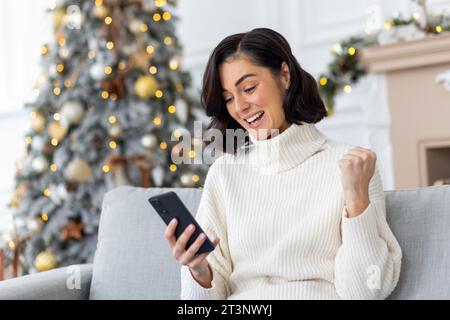 Happy young woman sitting at home on sofa near Christmas tree and using mobile phone, happy with success and good news for New Year holidays, showing victory gesture with hand. Stock Photo